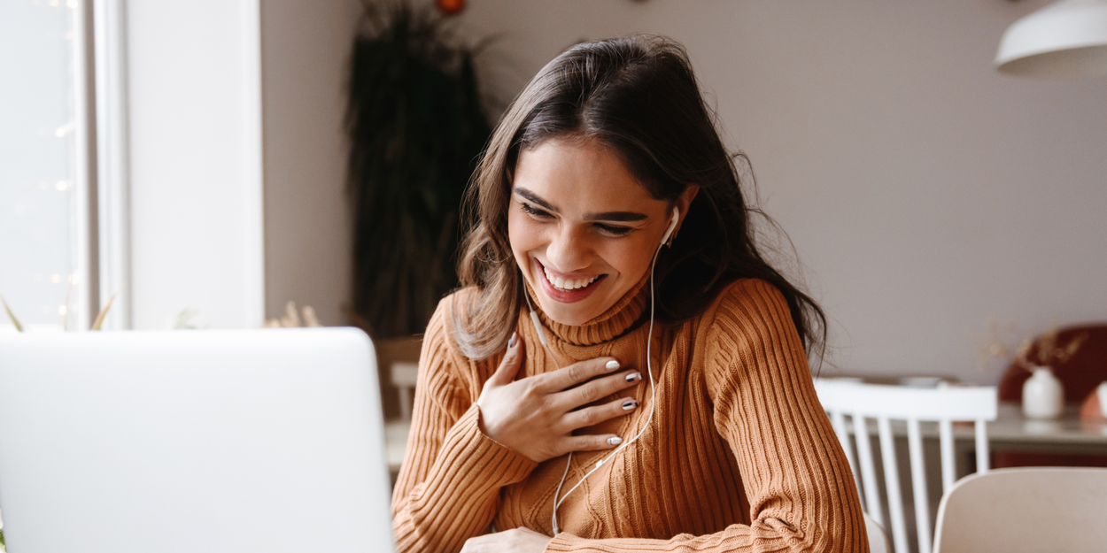 Woman in brown sweater laughing genuinely while talking to someone on laptop